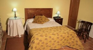 A bedroom with a wooden headboard, a bed with a patterned bedspread, two bedside tables with lamps, a chair, and a small table with a tablecloth. - Rectoral de Candas (Rairiz de Veiga)