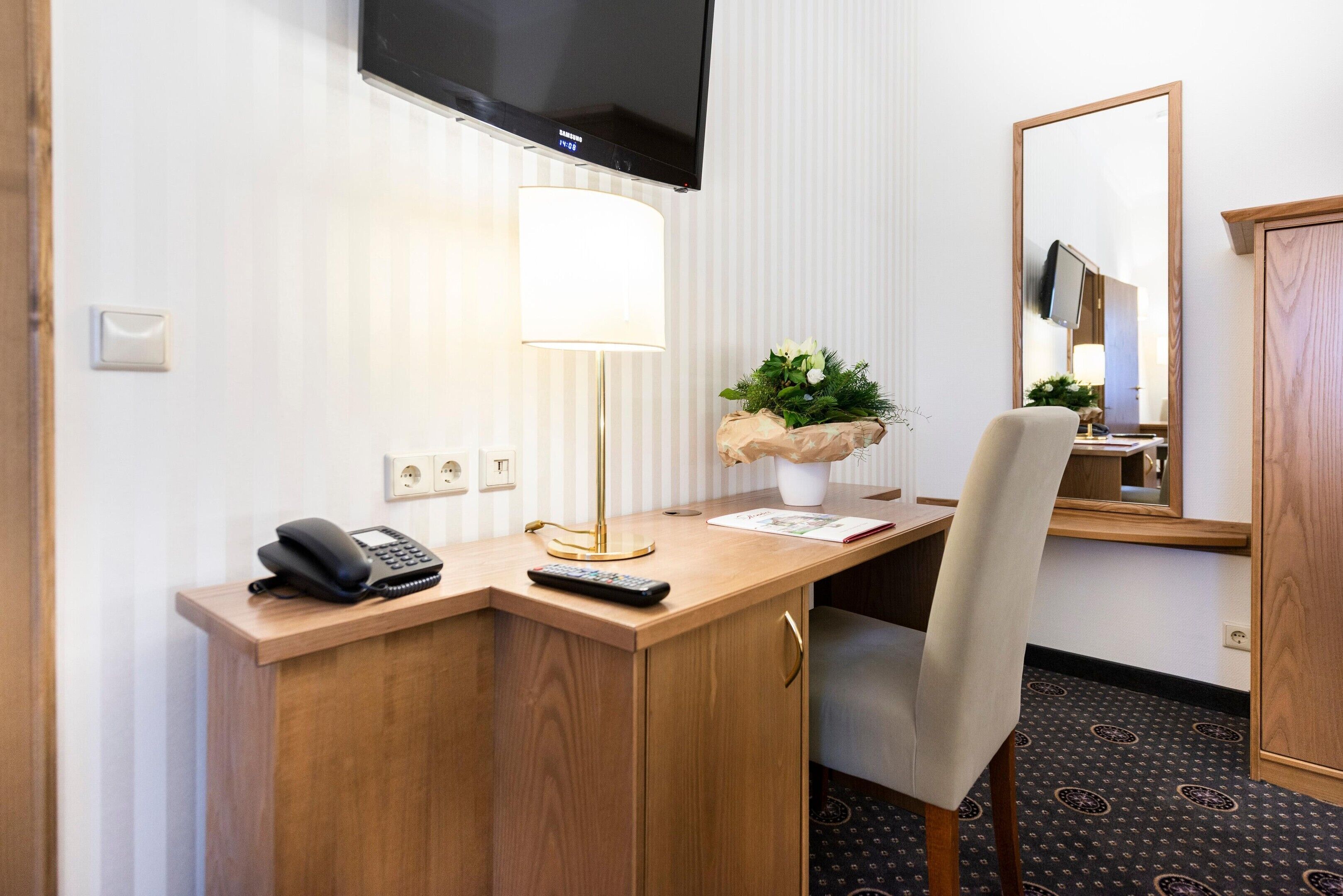 A hotel room with a wooden desk, a flat-screen TV, a lamp, a telephone, a remote control, a plant, a mirror, and a patterned carpet.