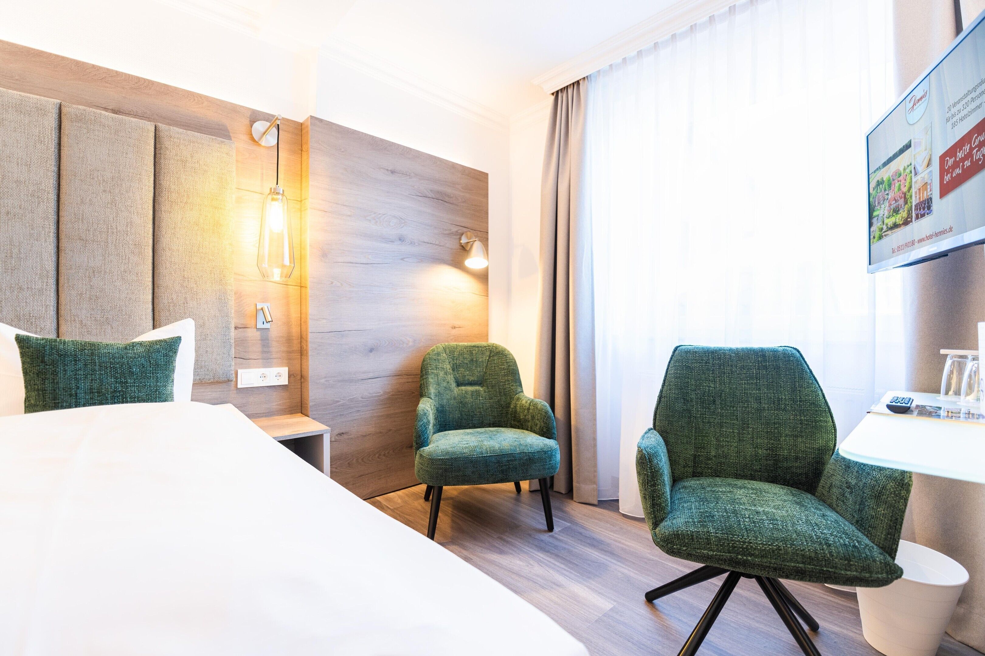 A hotel room with a bed, two green armchairs, a small table, a wall-mounted lamp, and a window with curtains.