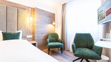 A hotel room with a bed, two green armchairs, a small table, a wall-mounted lamp, and a window with curtains.