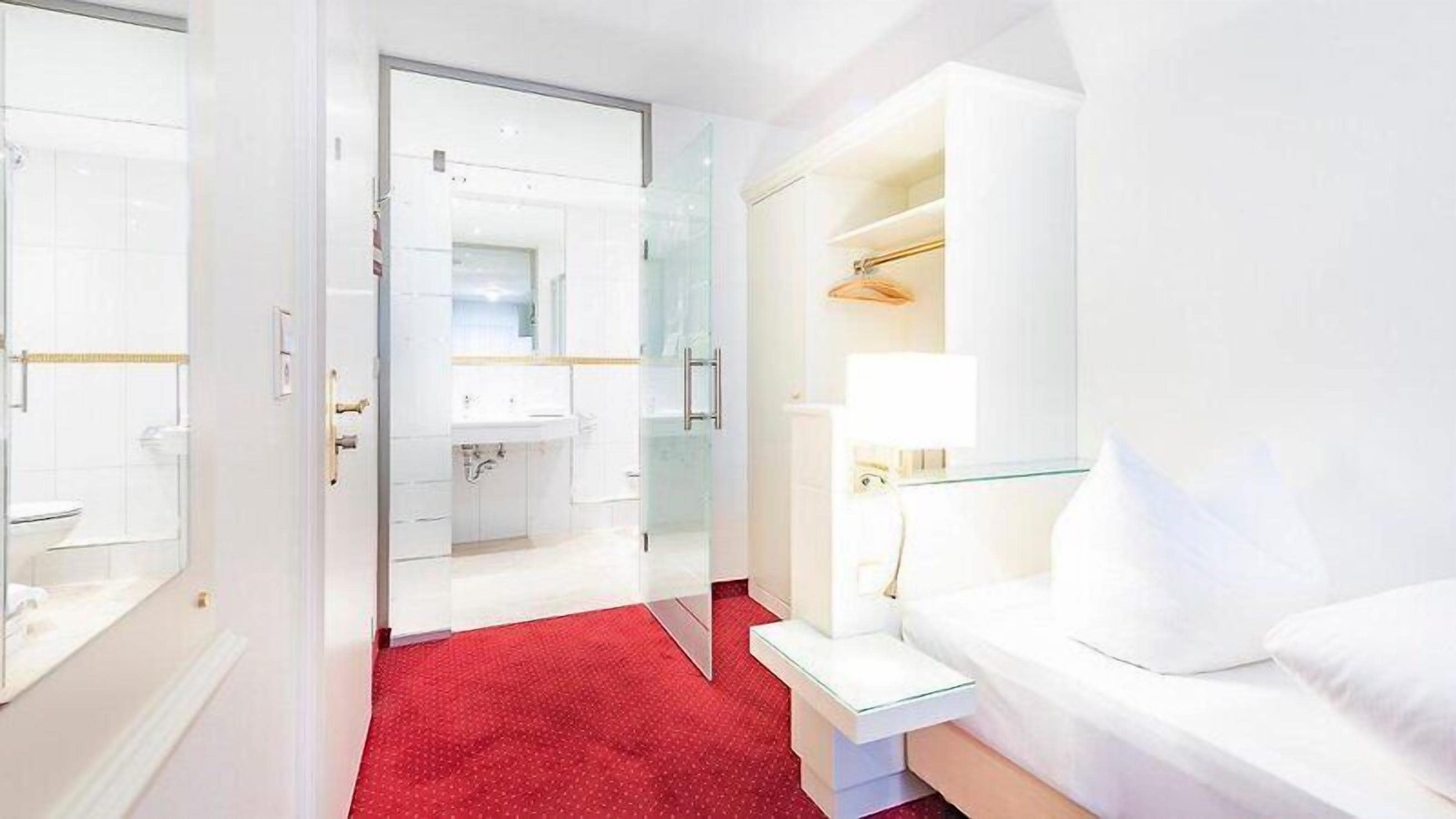 Quarto de hotel com carpete vermelho, cama branca, banheiro com paredes de vidro e guarda-roupa branco.
