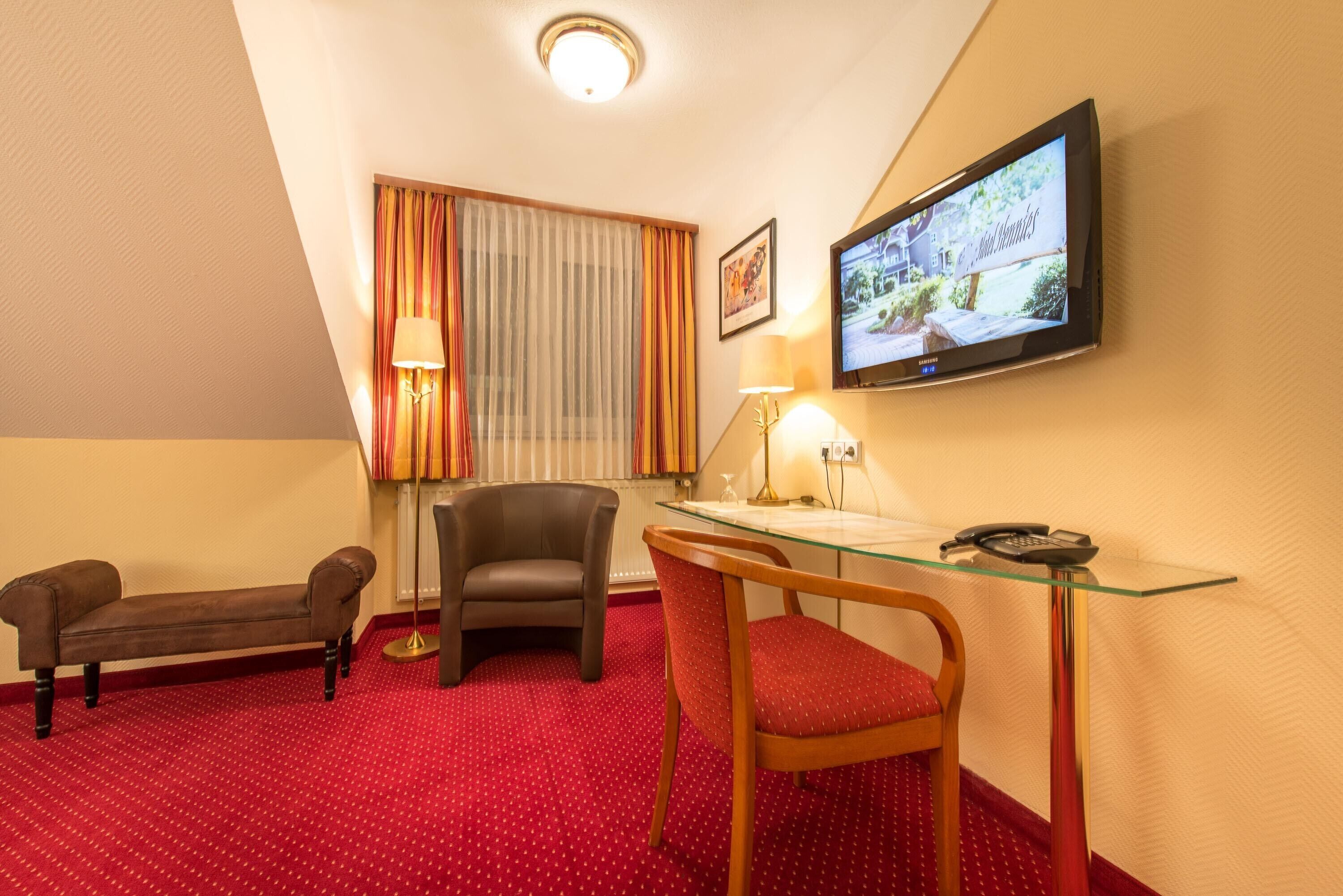 Habitación de hotel con alfombra roja, escritorio con teléfono, una silla, sofá, televisor montado en la pared y ventana con cortinas.