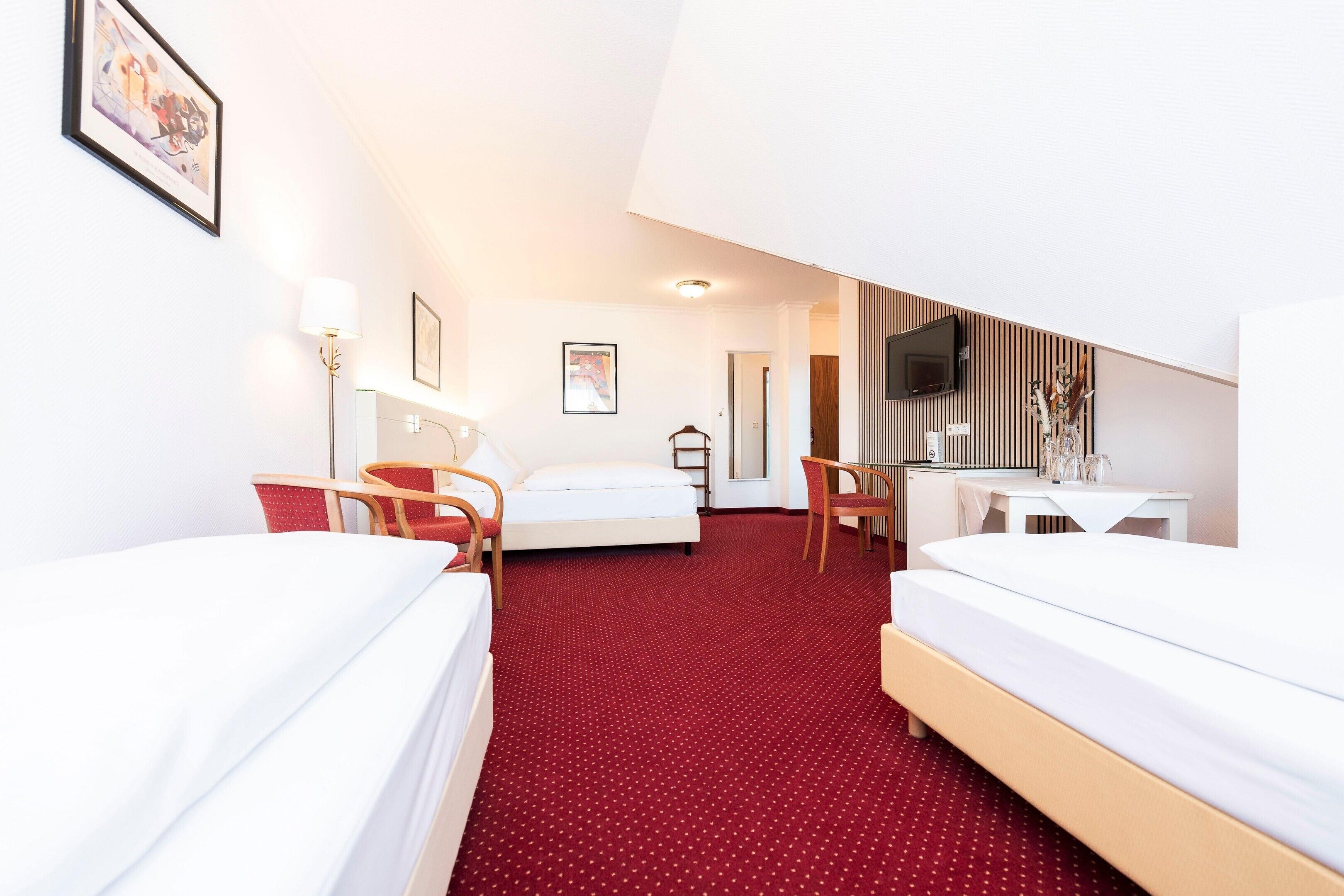 A hotel room with two beds, a desk, a chair, a TV, and artwork on the walls.
