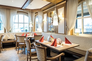 A restaurant with a long table, chairs, and a bench. There are curtains and windows with views. - Hotel Hennies (Isernhagen)