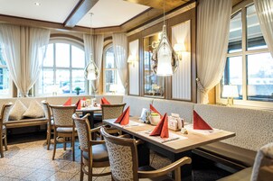 A restaurant with a long table, chairs, and a bench. There are curtains and windows with views.