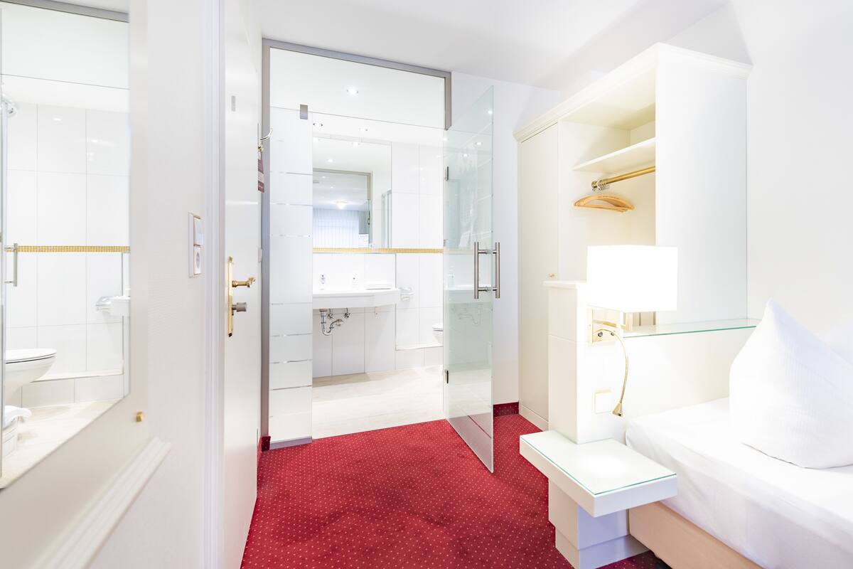 Hotellihuone, jossa on lasiseinällä varustettu suihku, valkoinen sänky, yöpöytä lampun kanssa ja punainen matto.