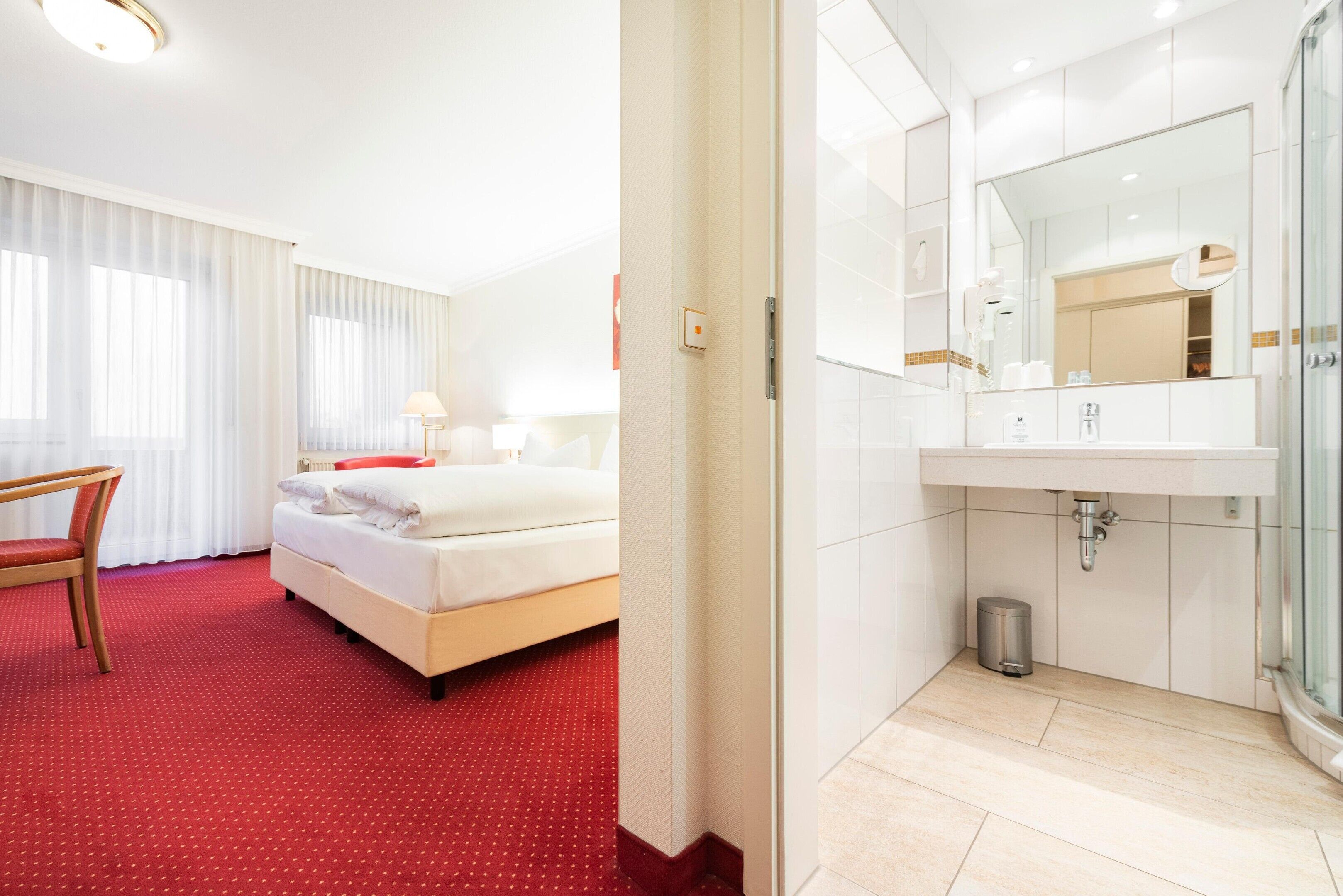 A hotel room with a bed, a chair, a window with curtains, a bathroom with a sink, mirror, and toilet.