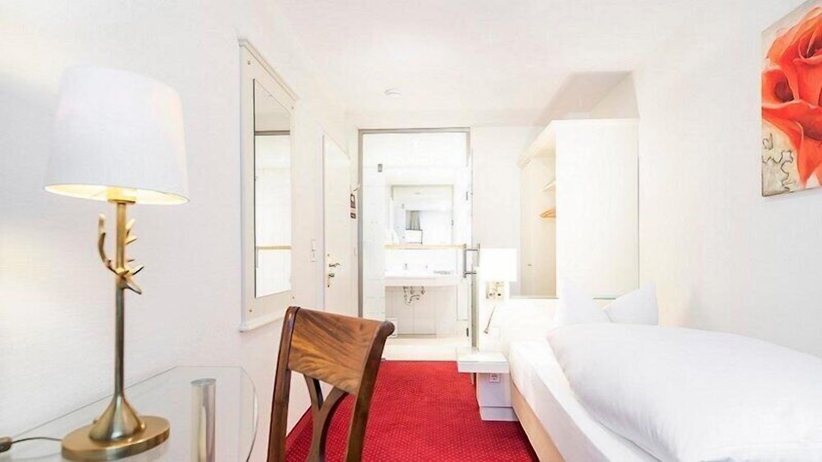 Une chambre d’hôtel avec un lit, une chaise, un bureau avec une lampe et une moquette rouge.
