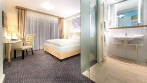 A hotel room with a bed, a desk with a lamp, a chair, a large window with curtains, and a bathroom with a sink and mirror. - Hotel Hennies (Isernhagen)