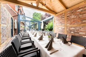 A restaurant with a long table set for dining, wicker chairs, and a brick wall. - Hotel Hennies (Isernhagen)