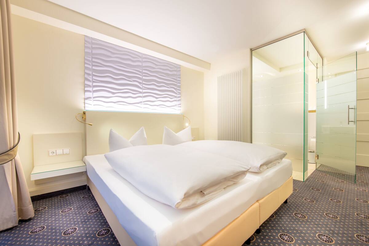 Hotellihuone, jossa on sänky, yöpöytä, aaltoilevakuviollinen ikkuna, lasinen suihkuseinä ja kuvioitu matto.