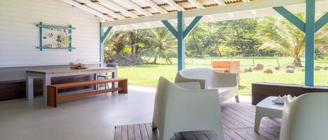 Un patio techado al aire libre con un banco de madera, sillas blancas y una mesa.