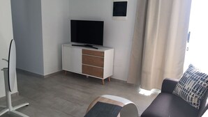 A modern living room with a flat-screen TV on a white cabinet, a sofa, and a small table with a rug. - Home from home 2 bed luxury apartment, heated pool. 202 (Puerto Del Carmen)