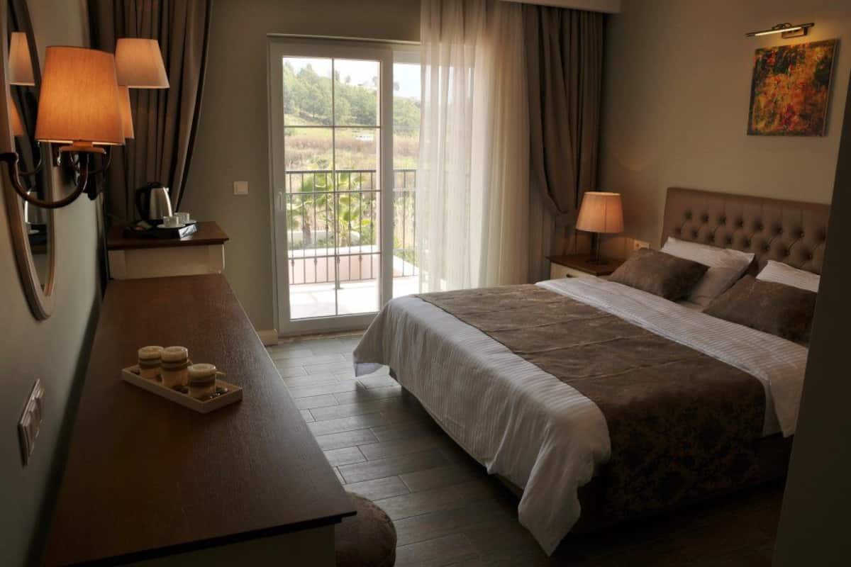 A hotel room with a large bed, two bedside tables with lamps, a wooden desk with a tray of items, and a balcony with a view of greenery.