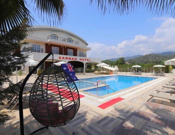 Zona de piscina con sillas de descanso y sombrillas, una silla colgante con una bandera, y un edificio con la leyenda 'SARBAN HOTEL' al fondo.