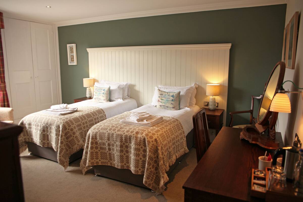 A hotel room with two beds, a wooden nightstand, a chair, and a desk with a lamp.