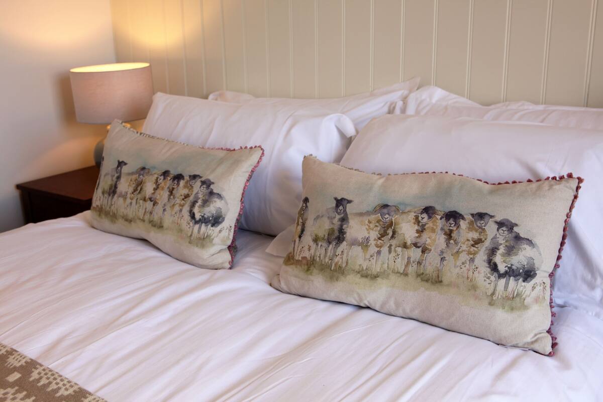 A bed with white linens and two decorative pillows featuring a pastoral scene.