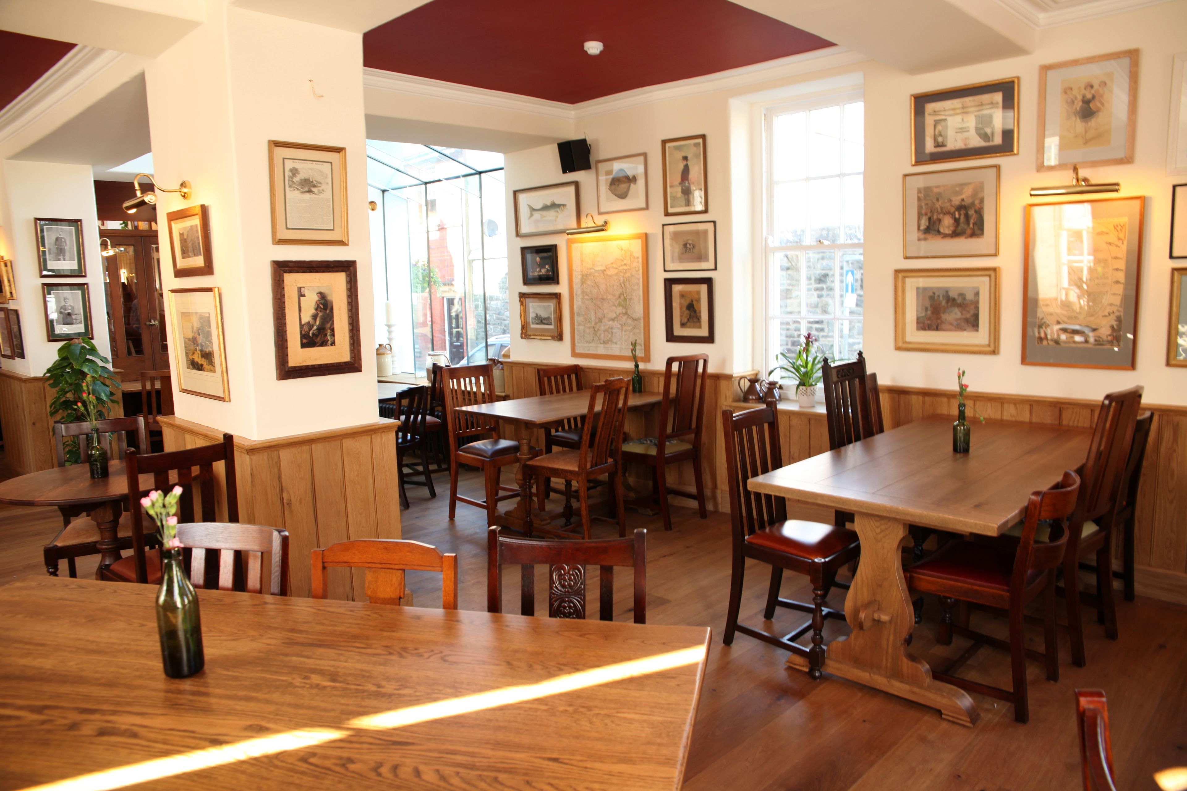 A restaurant interior with wooden tables and chairs, framed pictures on the walls, and a large window providing natural light.