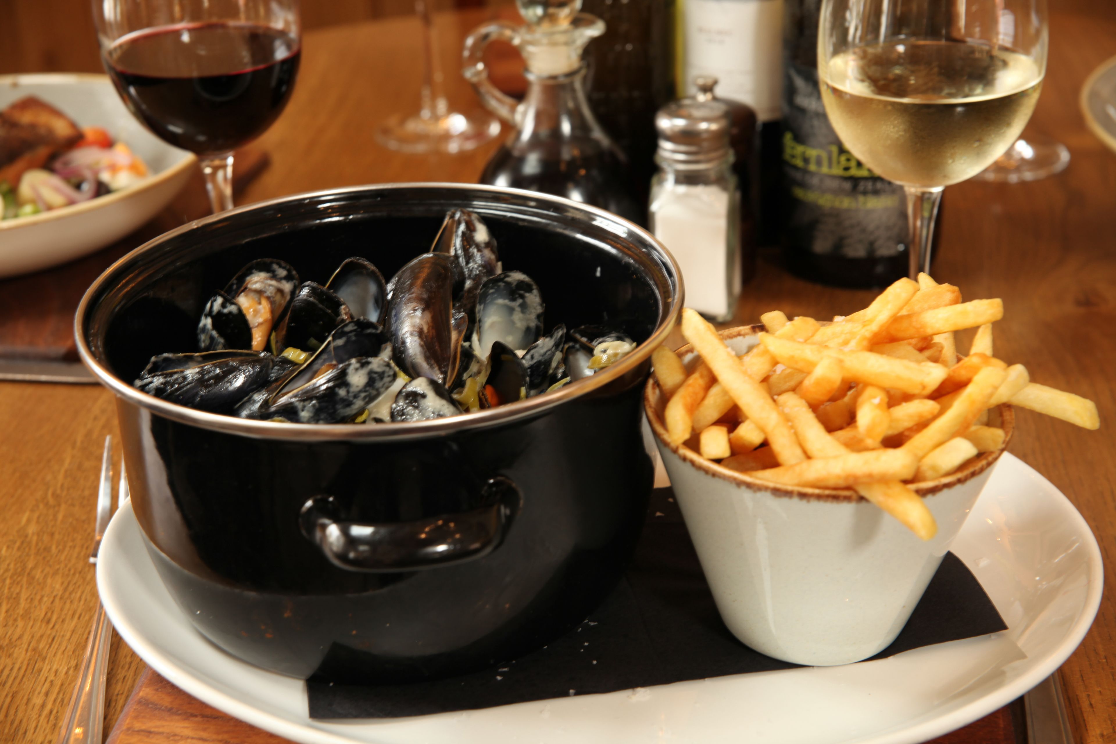 A dining table with a bowl of mussels, a cup of fries, and two glasses of wine.