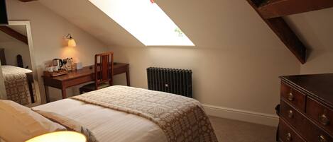 A bedroom with a sloped ceiling, a bed with a patterned bedspread, a dresser, a desk with a lamp, and a radiator.