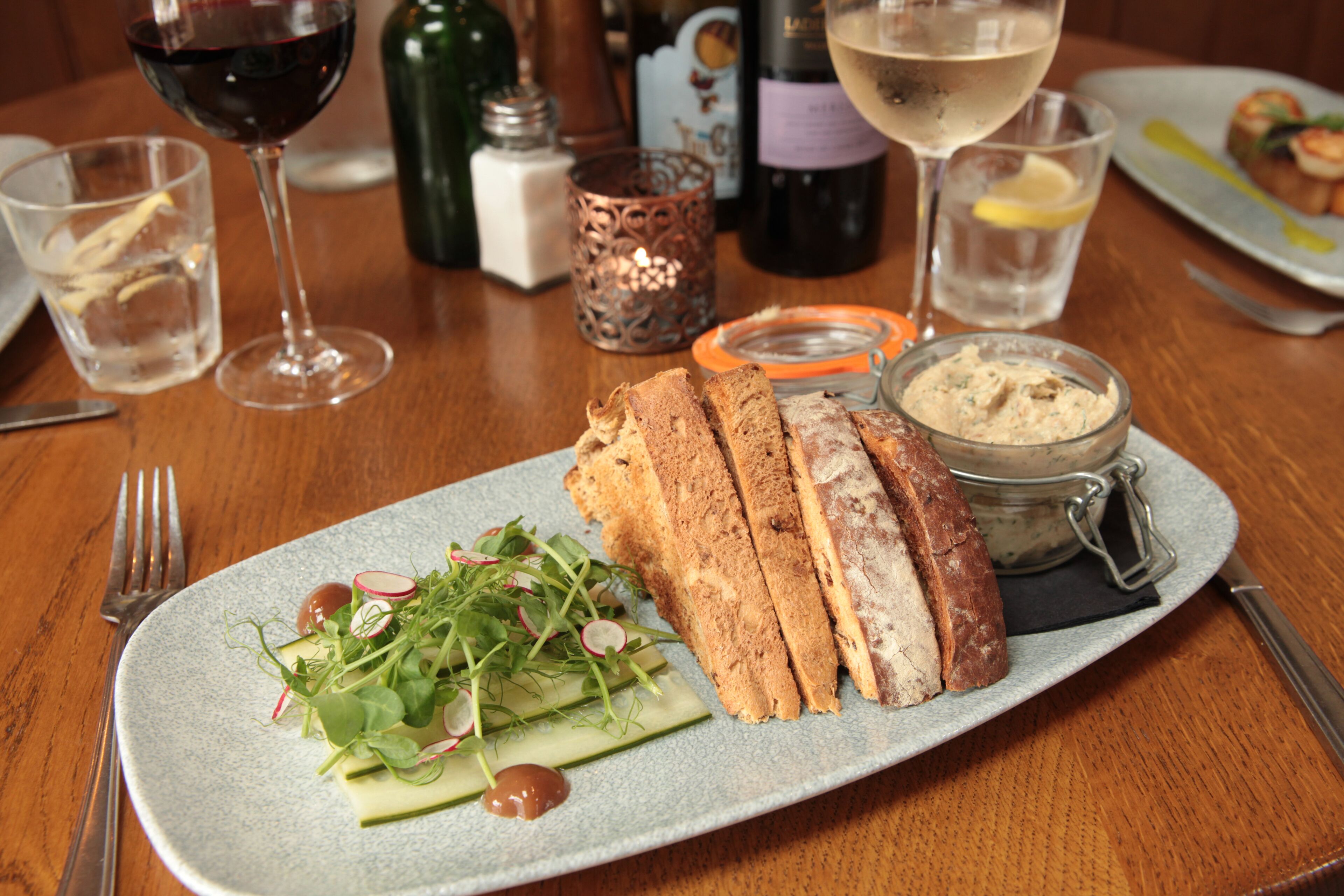 Un plato con pan en rebanadas, una ensalada y un frasco de crema sobre una mesa con copas y botellas de vino.