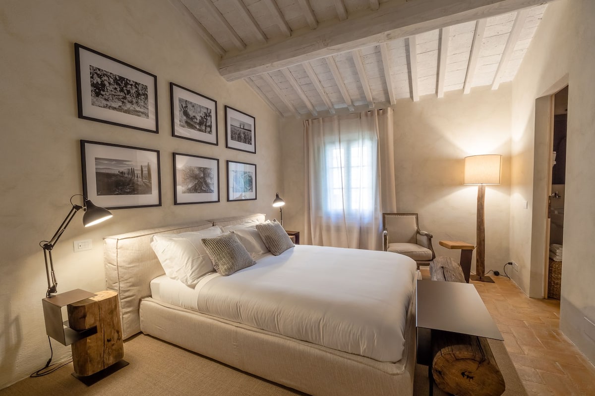 A bedroom with a large bed, a wooden nightstand, a desk, and a chair. There are framed pictures on the wall and a window with curtains.
