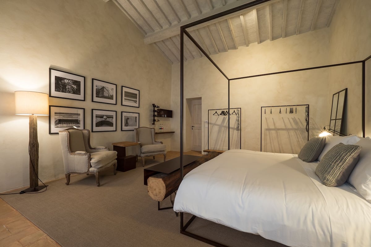 A bedroom with a four-poster bed, a wooden nightstand, a chair, a lamp, and framed pictures on the wall.