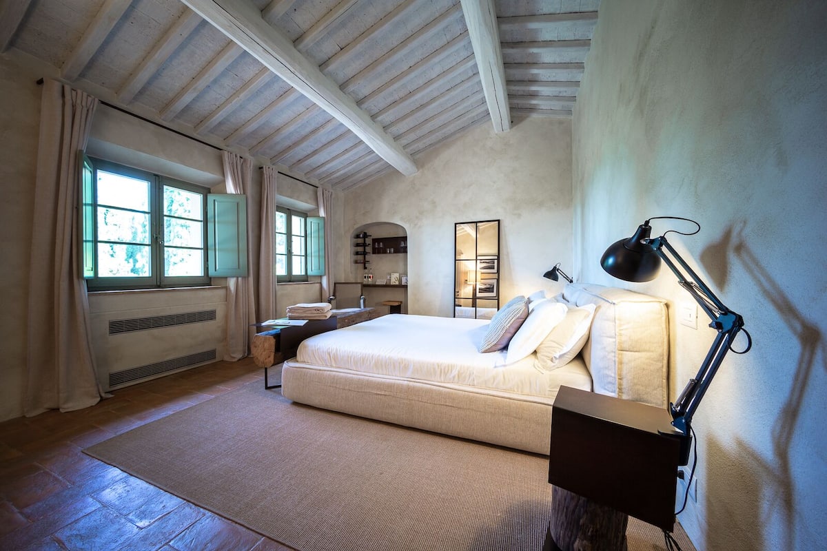 A bedroom with a large bed, a desk, a chair, a lamp, and a window with curtains.