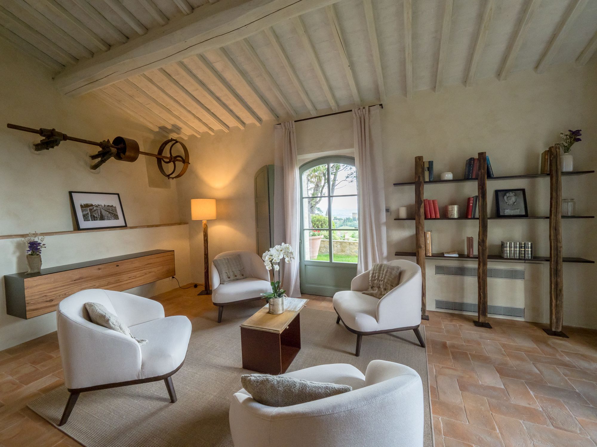 A spacious living room with a high ceiling, a wooden bench, two white armchairs, a coffee table, and a bookshelf.