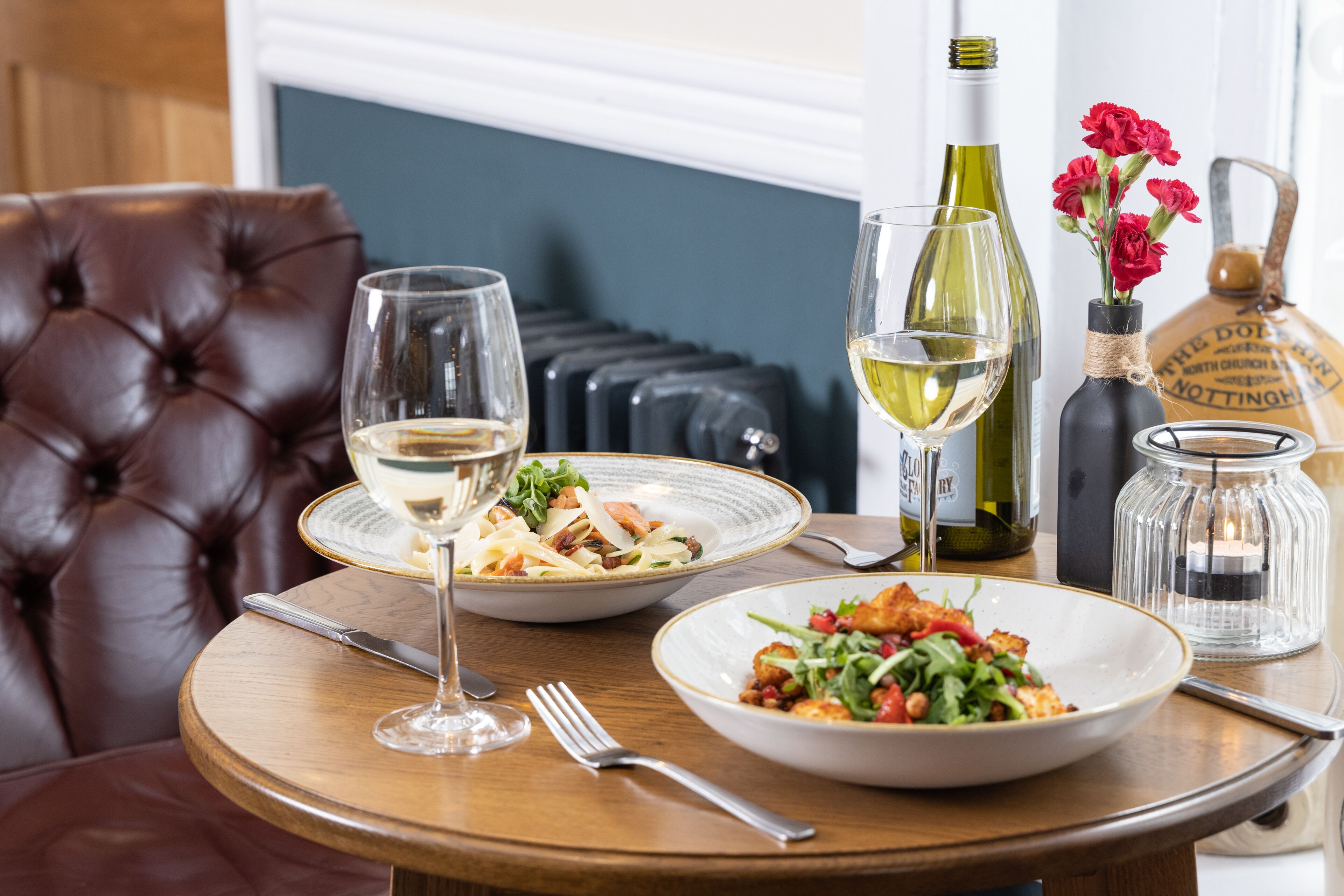A dining table with two plates of food, two wine glasses, a bottle of wine, a candle, and a vase with flowers.