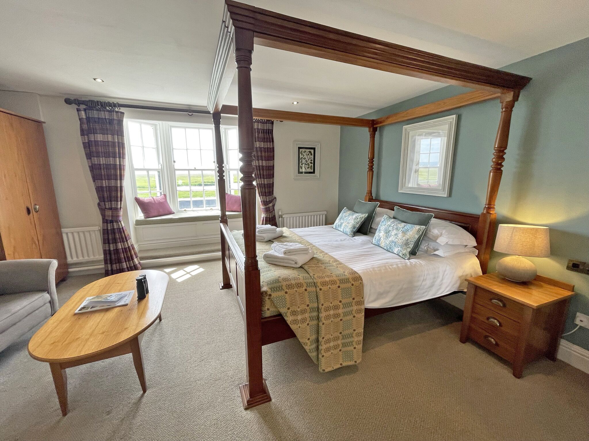 A four-poster bed with a wooden frame, a bedside table with a lamp, and a view of the outdoors through a window.