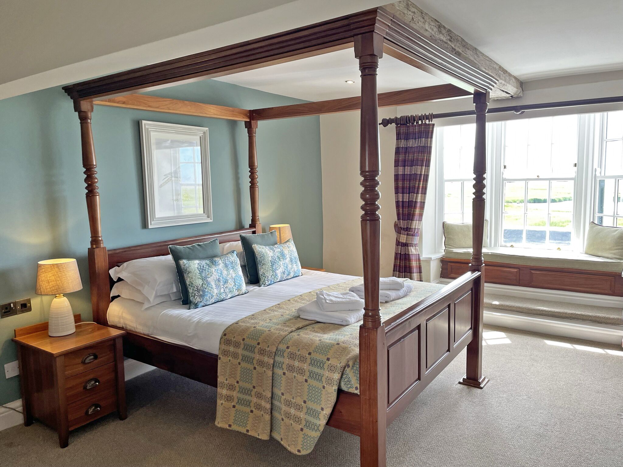 A four-poster bed with a wooden frame, a bedside table with drawers, and a window with a view of greenery.