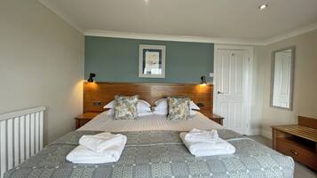 Een net opgemaakt bed met een geborduurd sprei en twee kussens, een nachtkastje met een lamp en een deur aan de rechterkant.