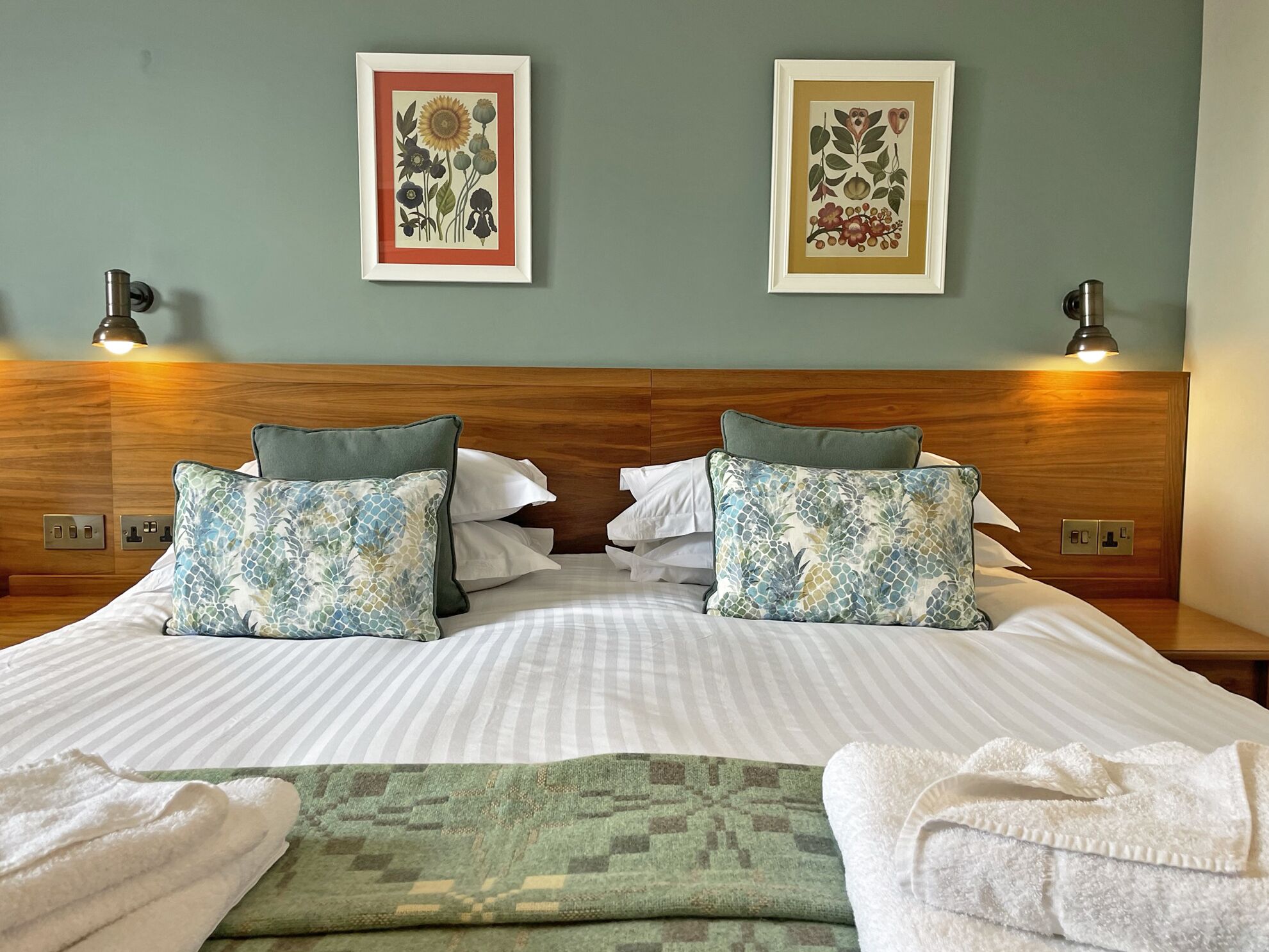 A neatly made bed with patterned pillows, two framed botanical prints on the wall, and a nightstand with a lamp.