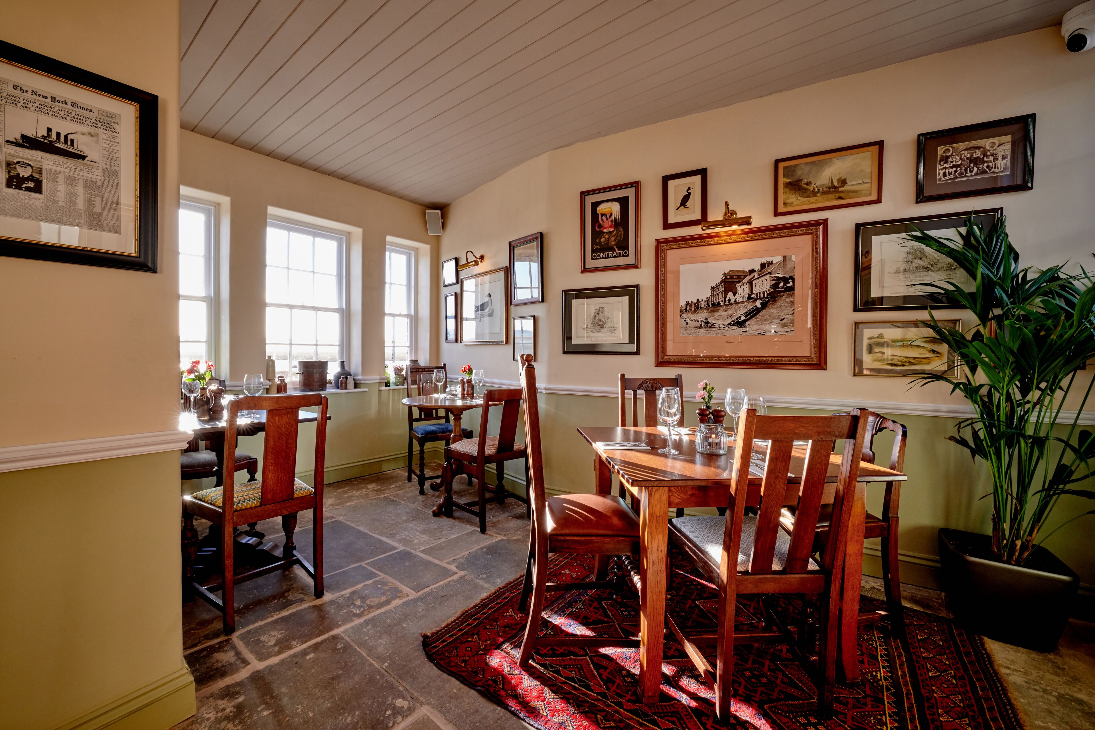 A dining area with a wooden table and chairs, framed pictures on the wall, and a potted plant.