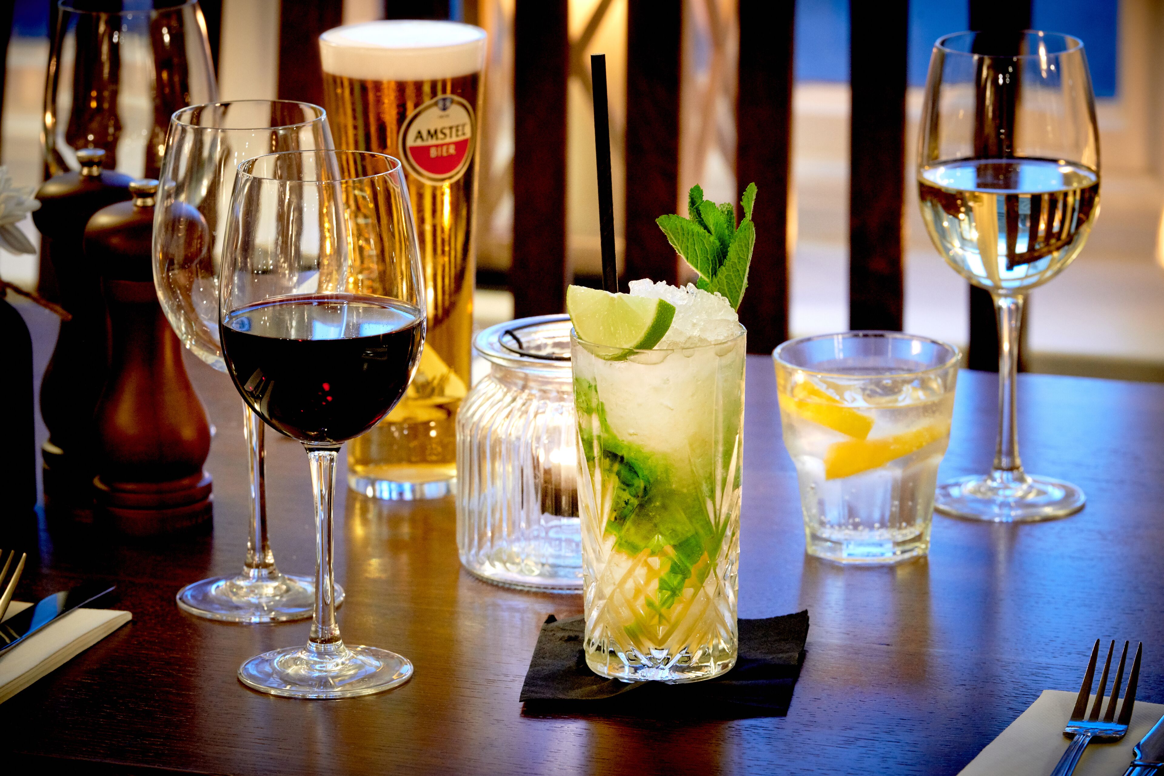A bar setting with various glasses of beverages, a beer mug, and a cocktail glass garnished with lime and mint.