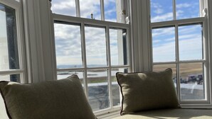Asiento junto a la ventana con dos cojines, vista a un cielo nublado y una pincelada de paisaje con carretera y vehículos.
