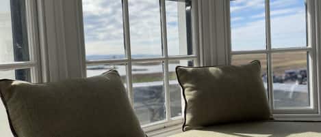 A window seat with two cushions, a view of a cloudy sky, and a glimpse of a landscape with a road and vehicles.