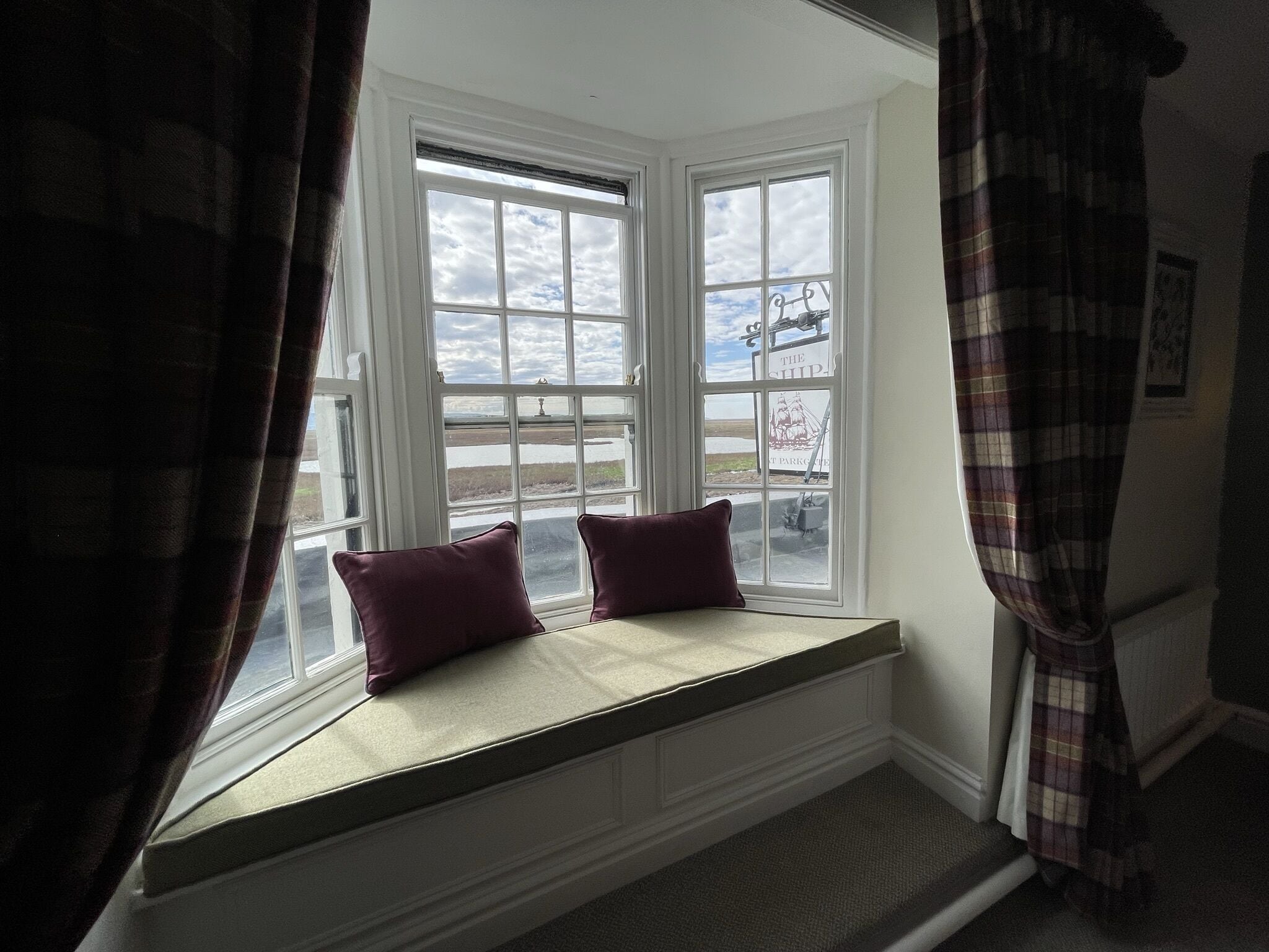 A bay window with two cushions, a view of a ship, and a plaid curtain.