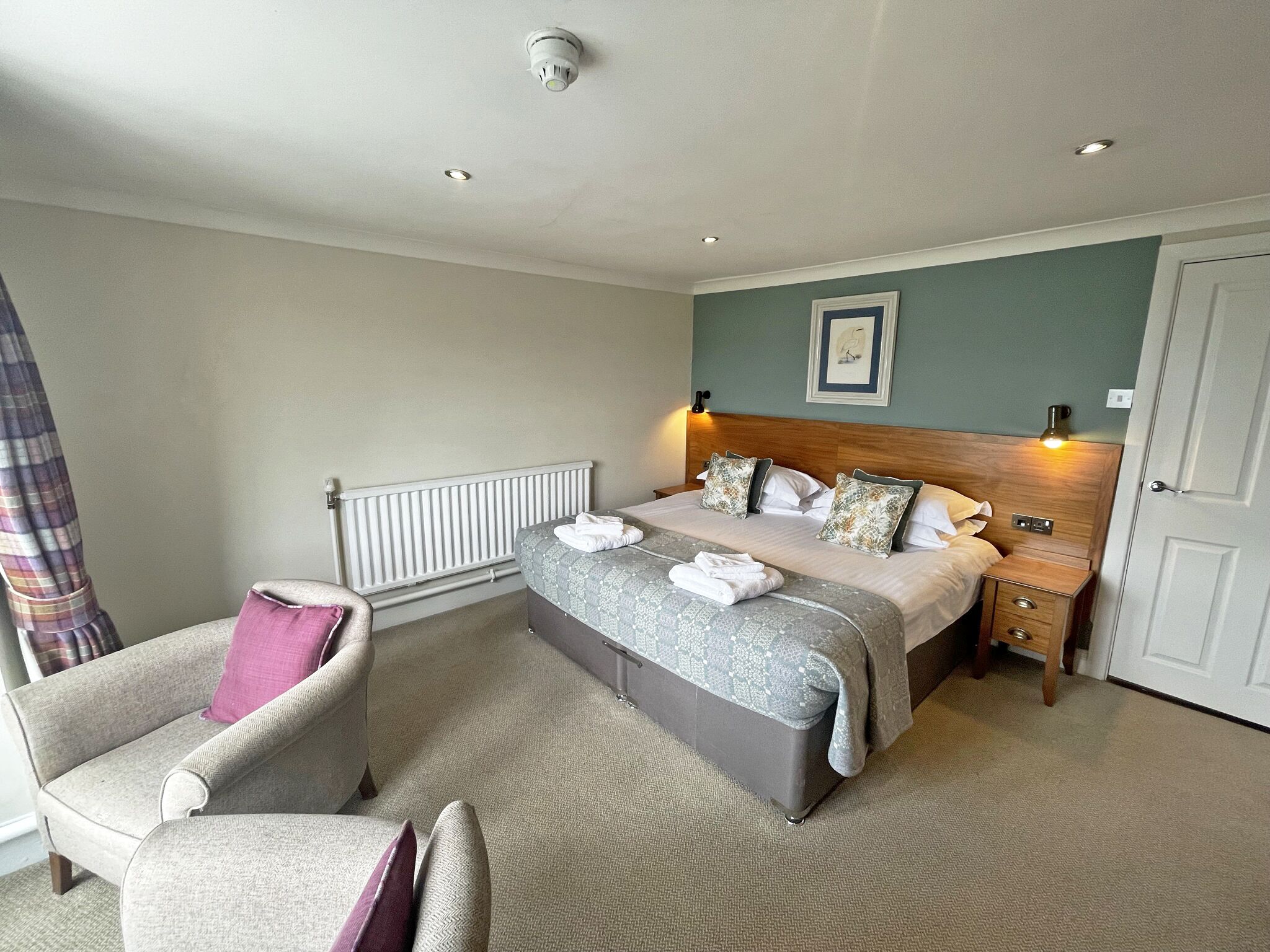 A hotel room with a bed, a chair, a bedside table, a radiator, and a door.