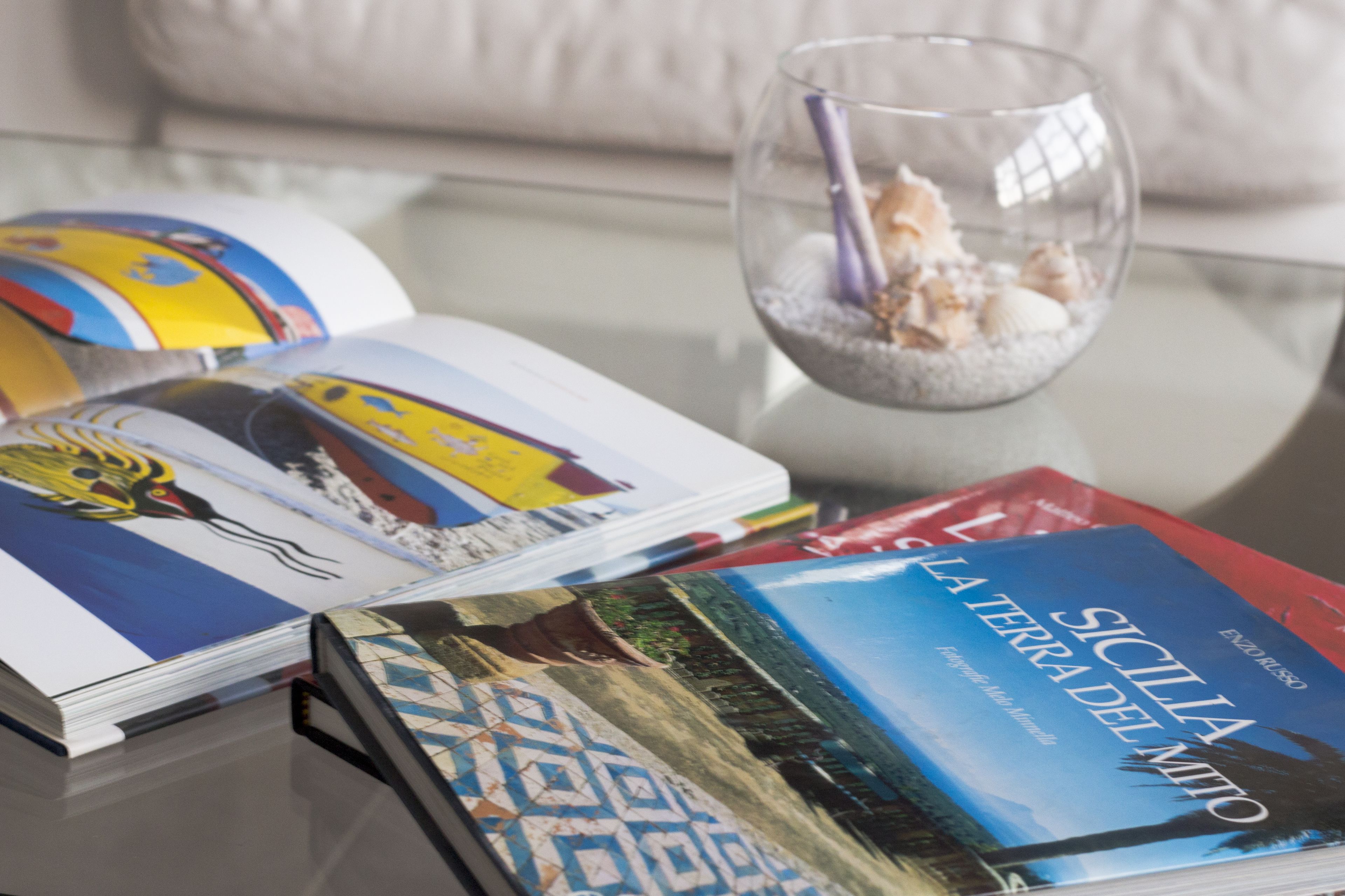 A glass table with a book titled "Sicilia La Terra Del Mito" and a glass bowl containing seashells and a purple straw.
