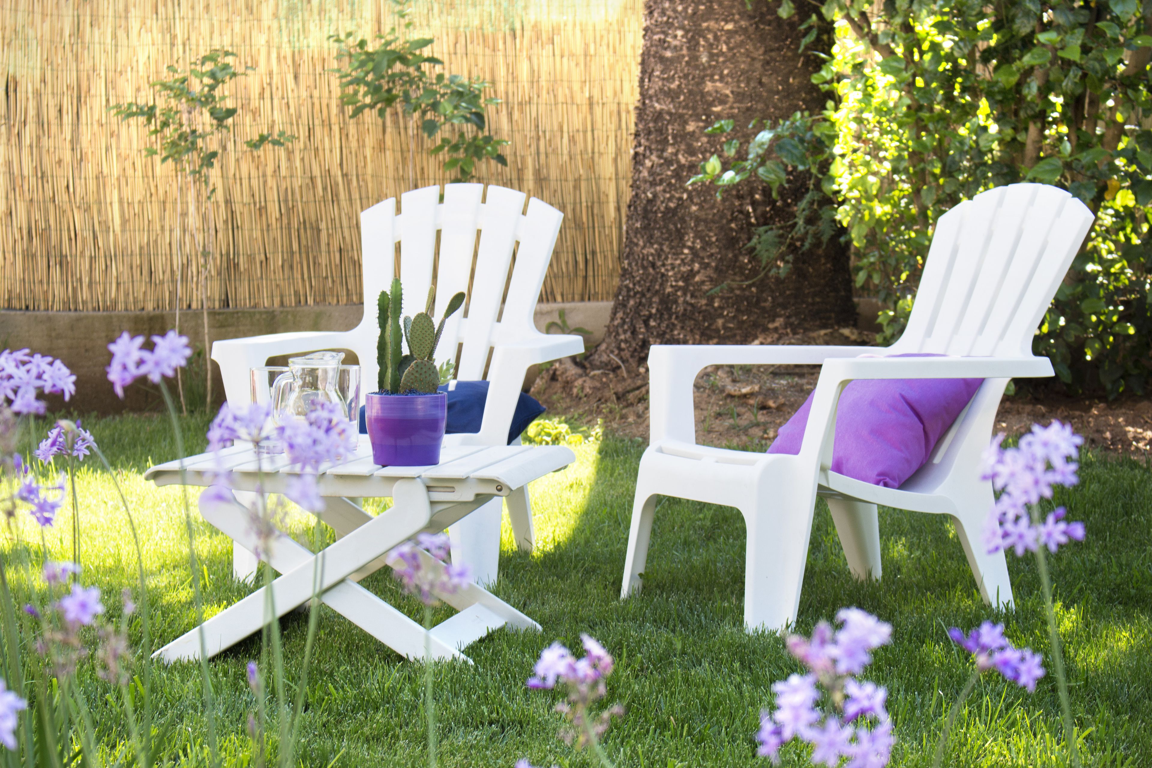 Two white chairs with purple cushions, a small table with a potted plant, and purple flowers on a grassy lawn.