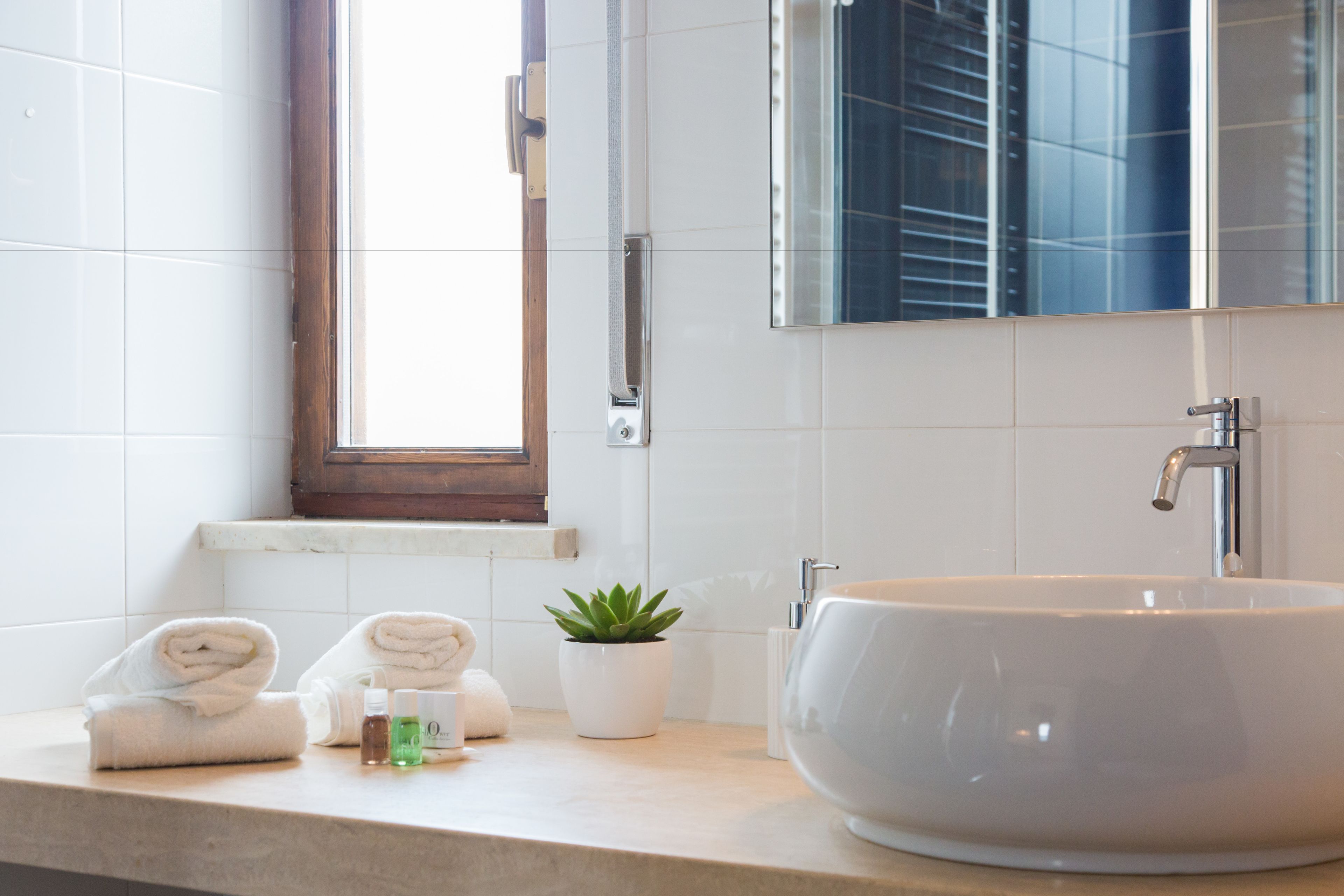 A bathroom with a large white sink, a mirror, a window, and a countertop with towels and a potted plant.
