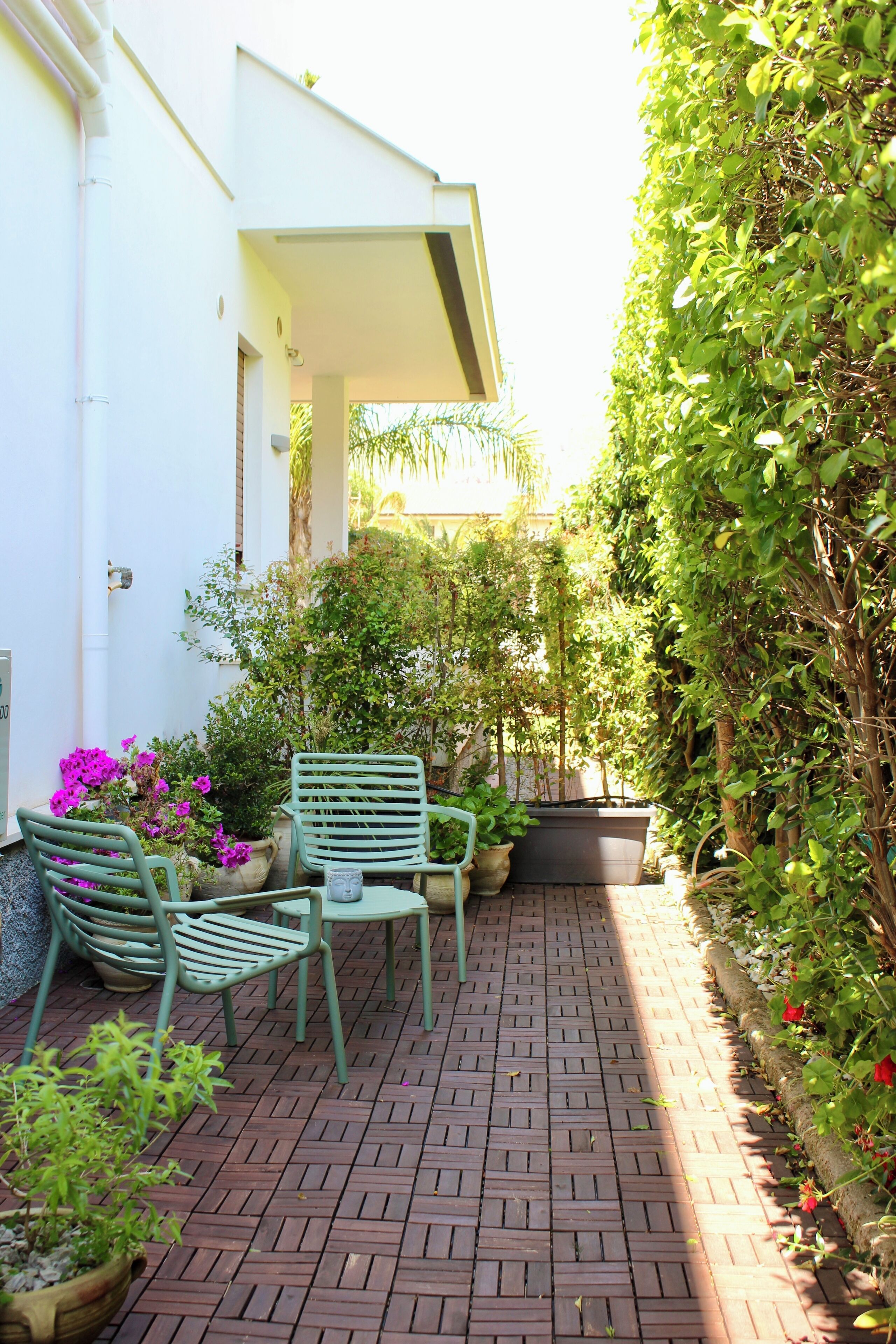 Une terrasse dallée avec deux chaises vertes et une table, entourée de plantes et de fleurs en pot.