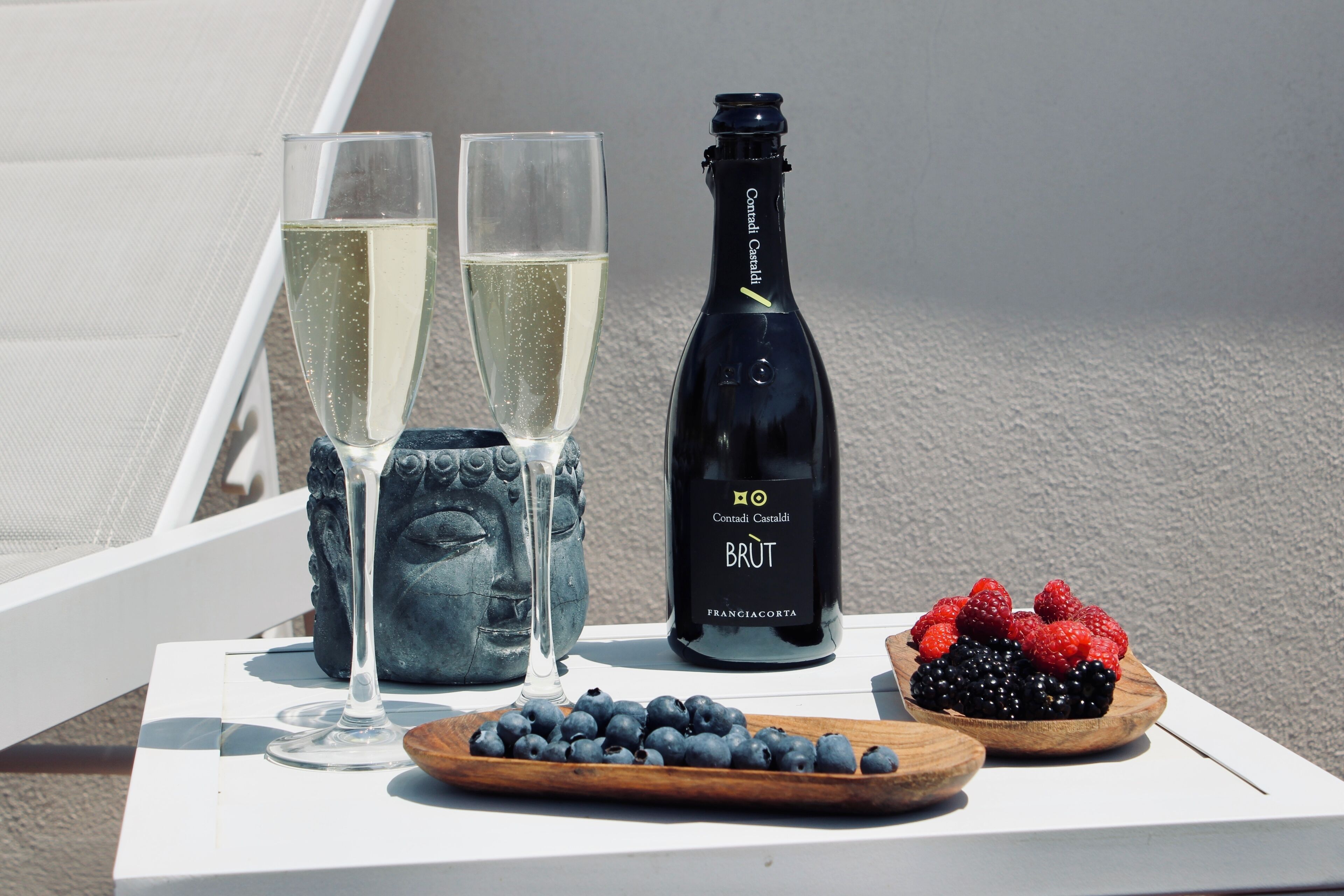 A table with two glasses of champagne, a bottle of Brut champagne, and a wooden board with blueberries and berries.
