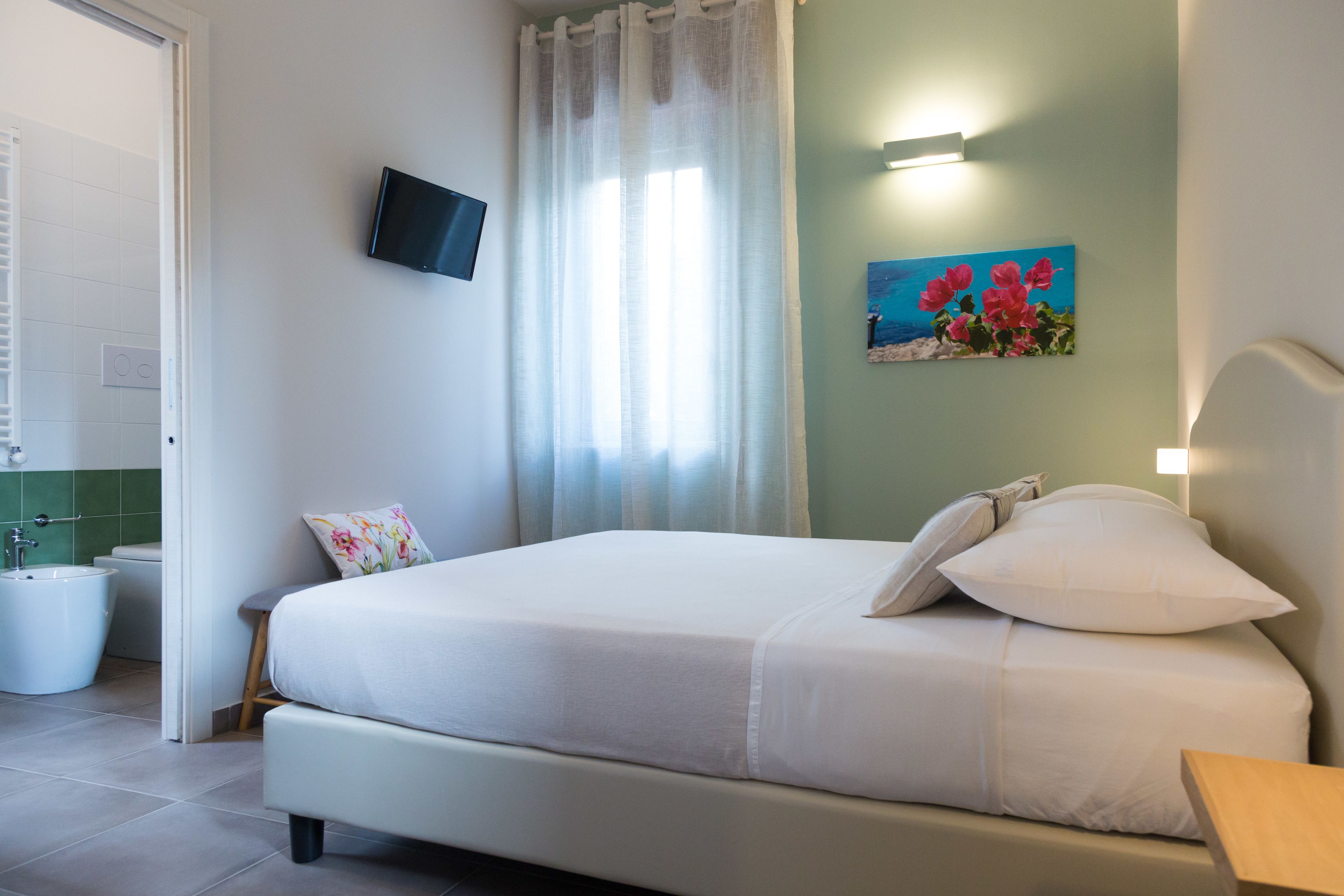 Una cama bien tendida con ropa de cama blanca, un televisor montado en la pared, un cuadro de flores y un baño con lavamanos e inodoro blancos.