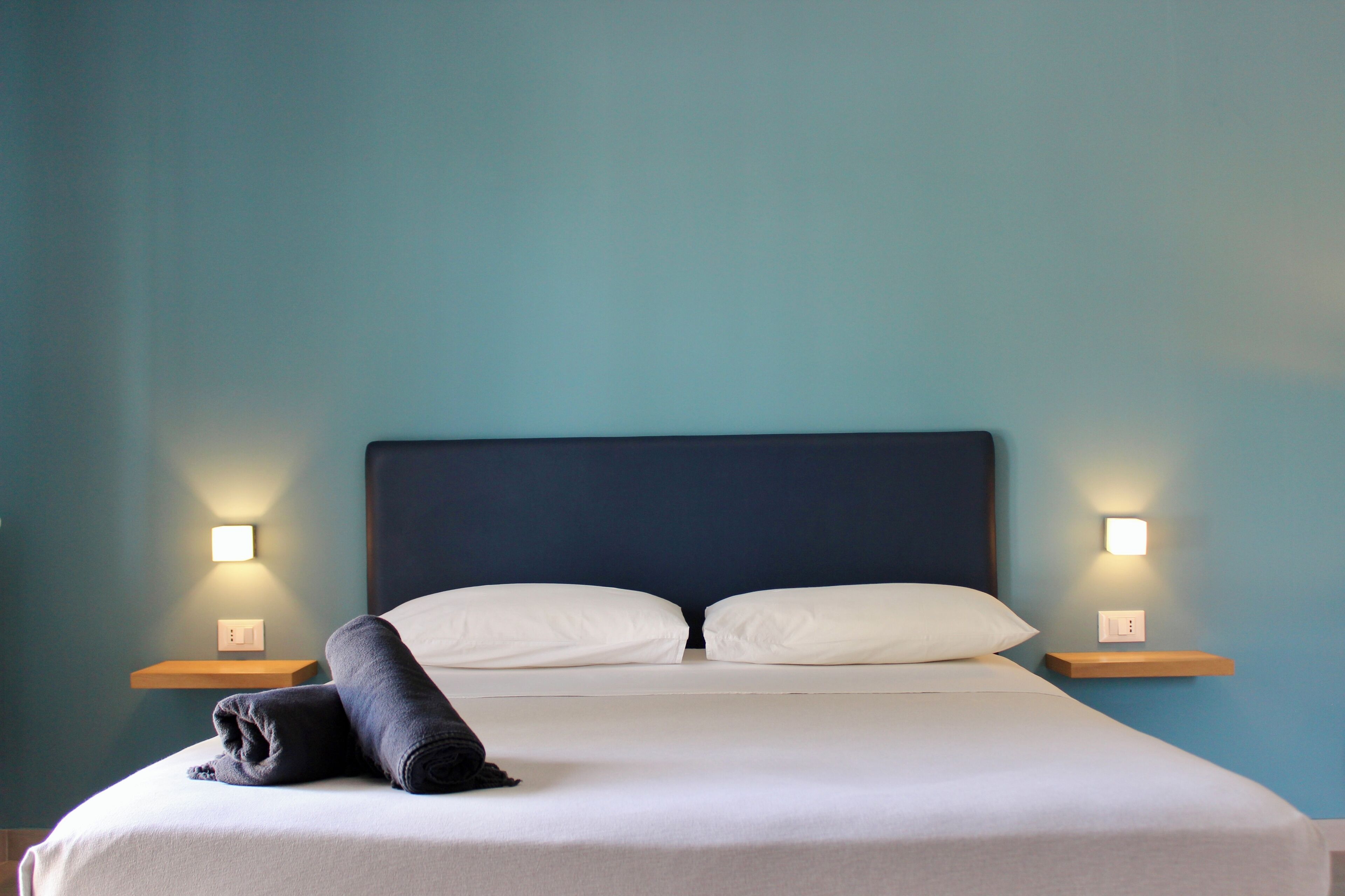 A neatly made bed with white linens and a rolled towel, two bedside tables with wall-mounted lamps, and a dark headboard.