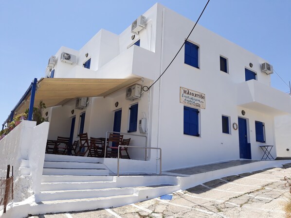 Un bâtiment blanc avec des portes et des fenêtres bleues, une enseigne et des sièges extérieurs avec des chaises.