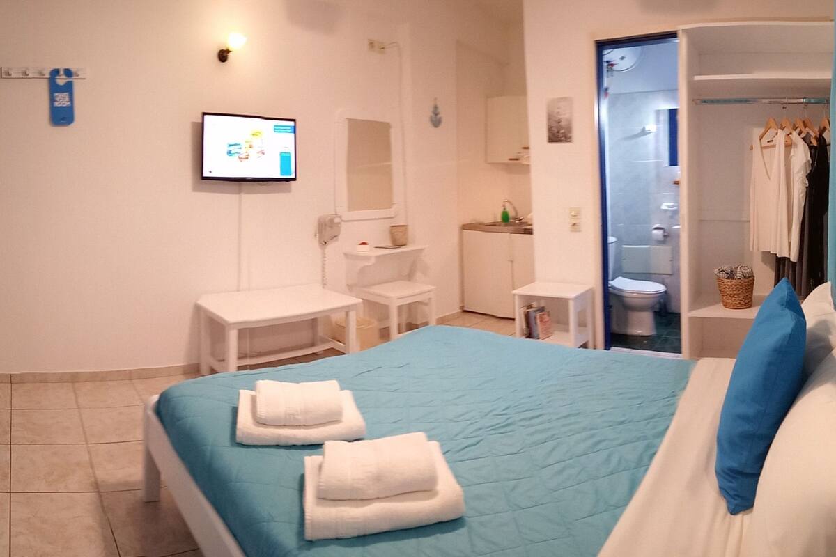 A hotel room with a bed, a desk, a chair, a TV, and a bathroom visible through an open door.