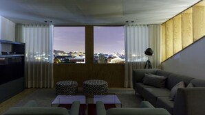 A modern living room with a large window, a sofa, two ottomans, a coffee table, and a tripod lamp. - Praça 66 Guest House (Porto)